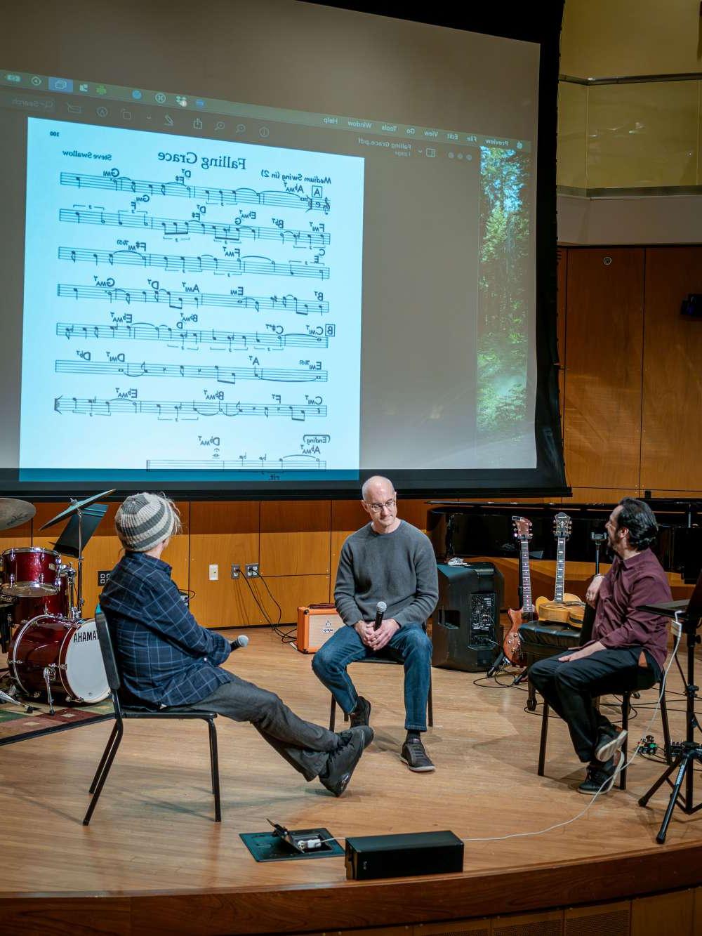 Three men onstage discuss a sheet of music projected overhead on a screen.