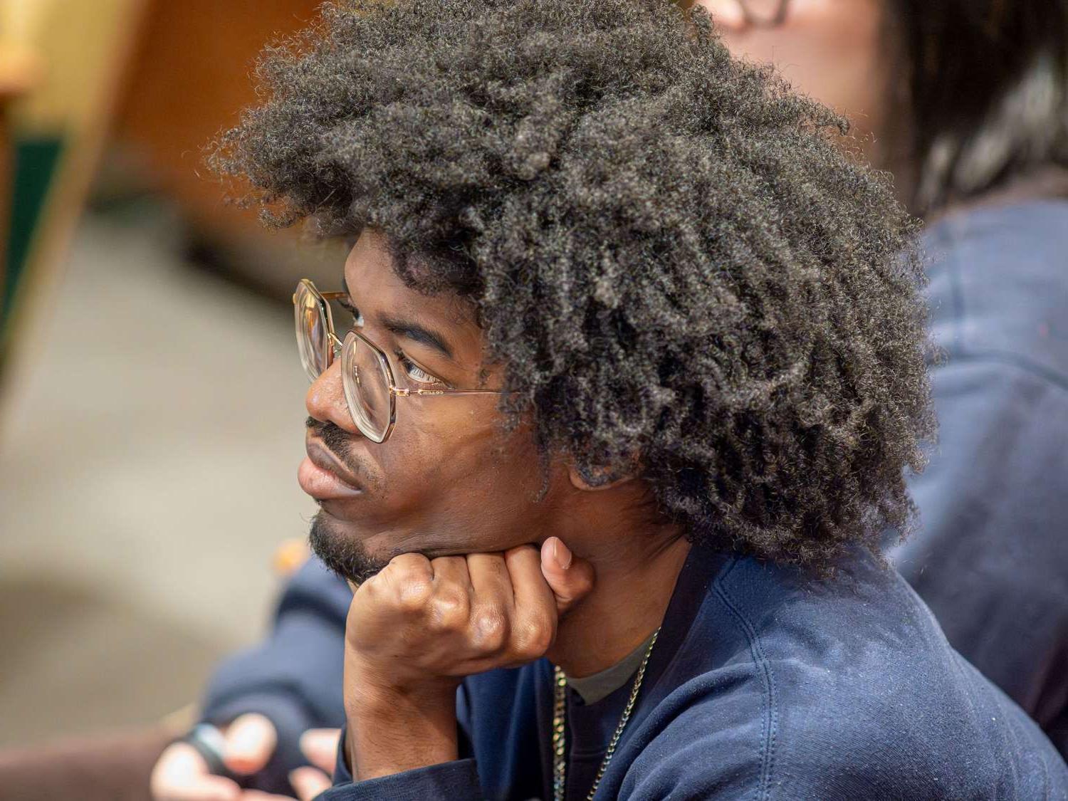 A student wearing glasses holds his hand under his chin in a class.